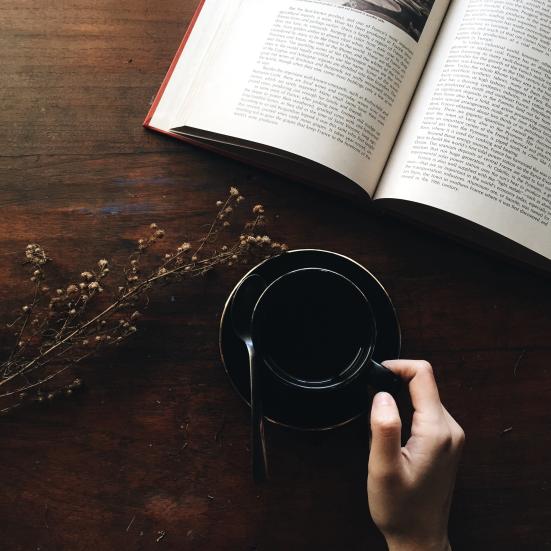En bog der ligger opslået på et bord og en hånd der holder en kop kaffe