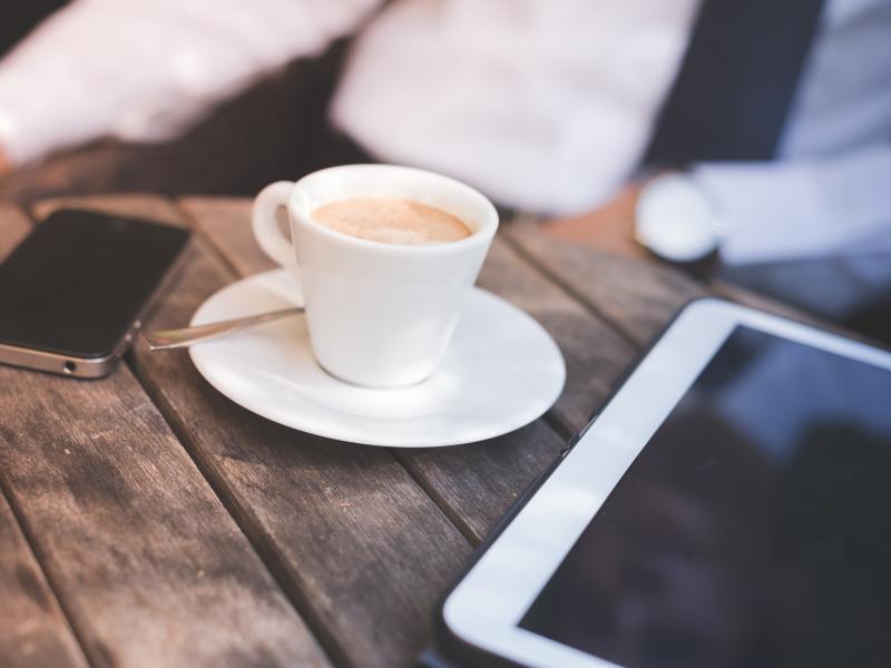 En tablet ligger på et bord og ved siden af står en kop kaffe